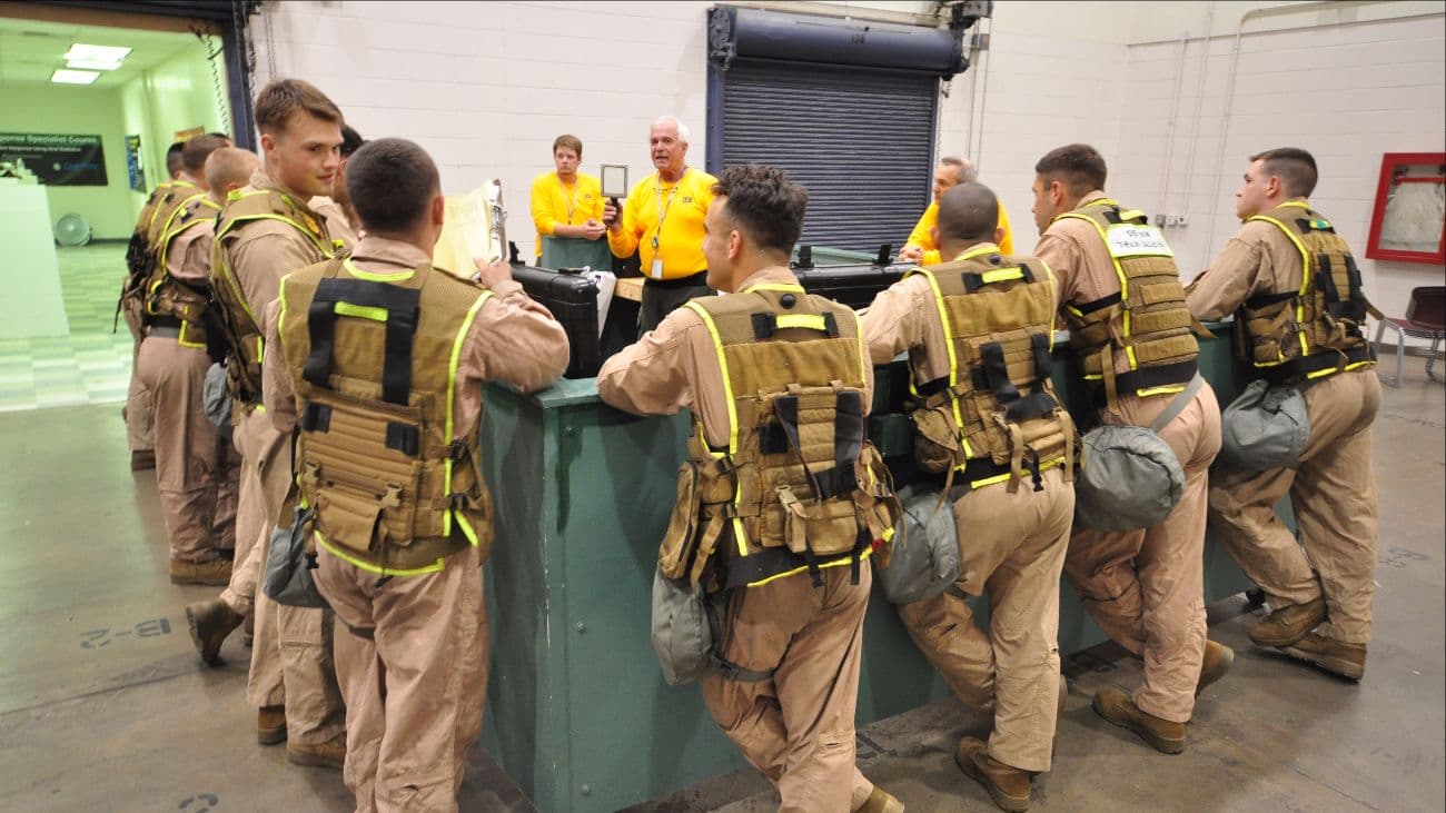 Marines in tan coveralls and gear gather around a table for a briefing inside a training bay.