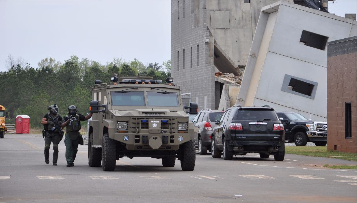 Two officers walk ahead of an armored tactical vehicle down a street of training buildings.