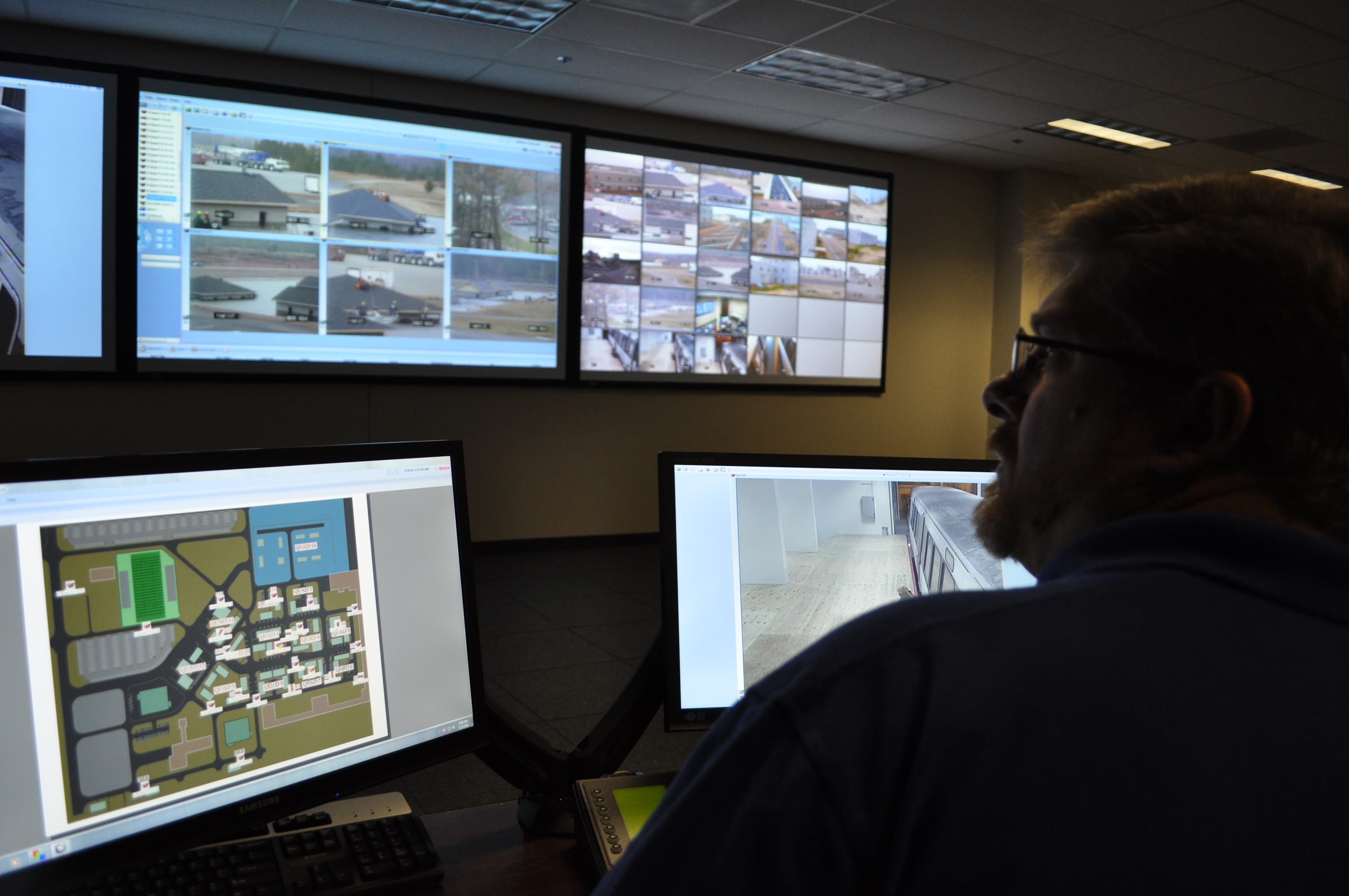An operator in the command center watches a wall of monitors showing camera feeds and a map of the campus.