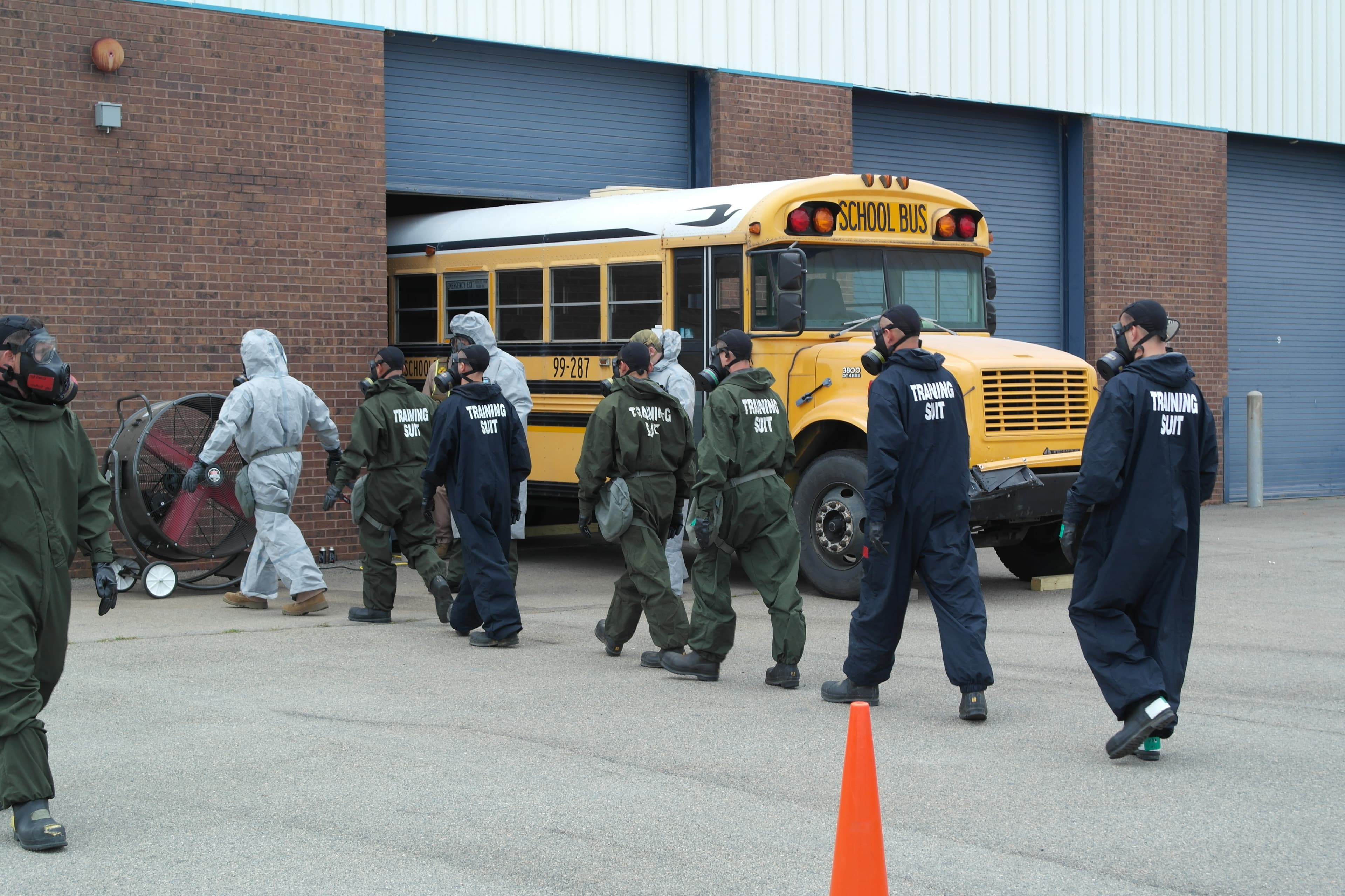 A training team in green protective suits and respirators walks past a school bus toward an open garage door.