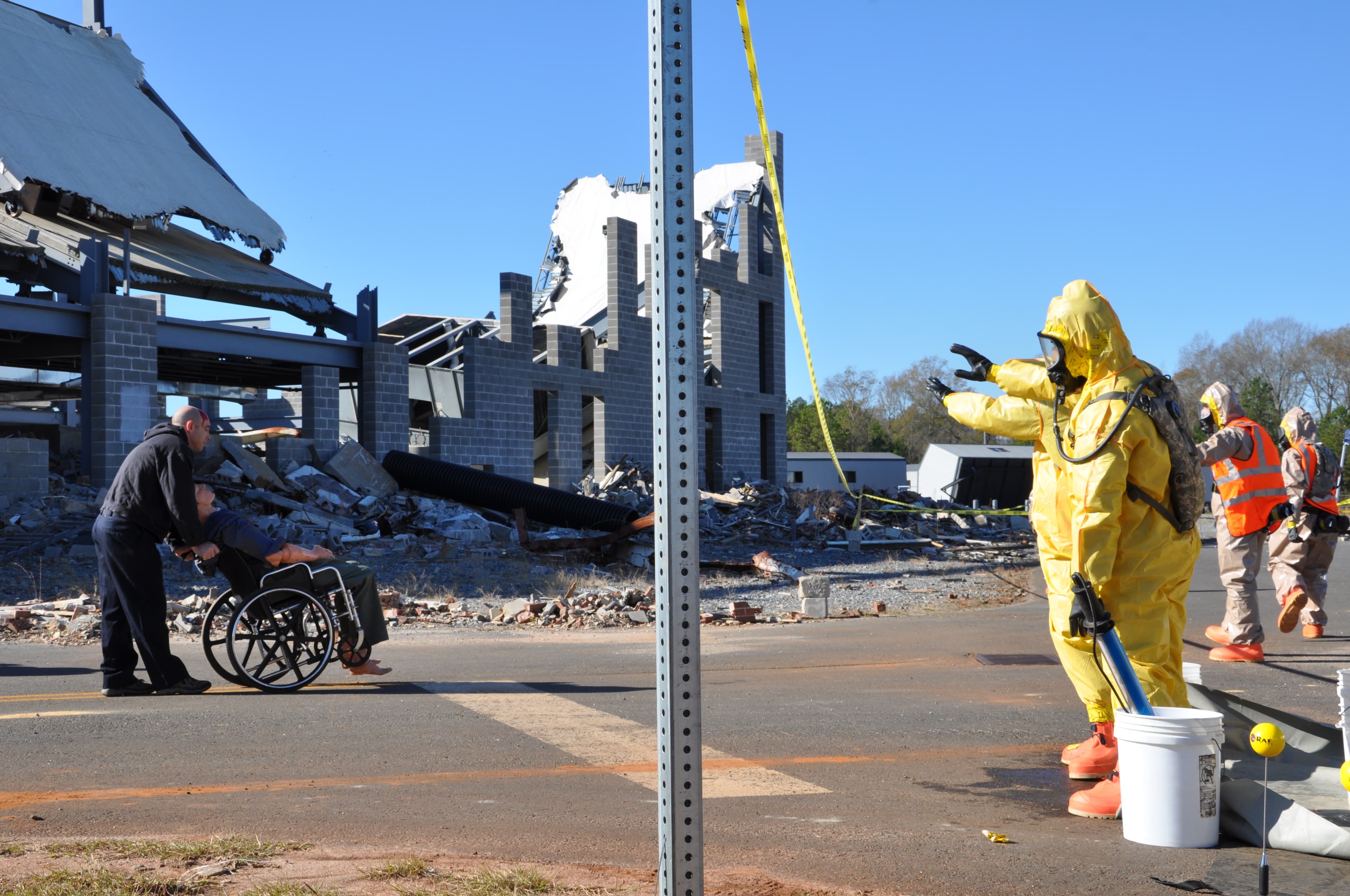 A responder in a yellow hazmat suit directs a man pushing a wheelchair away from a collapsed building.