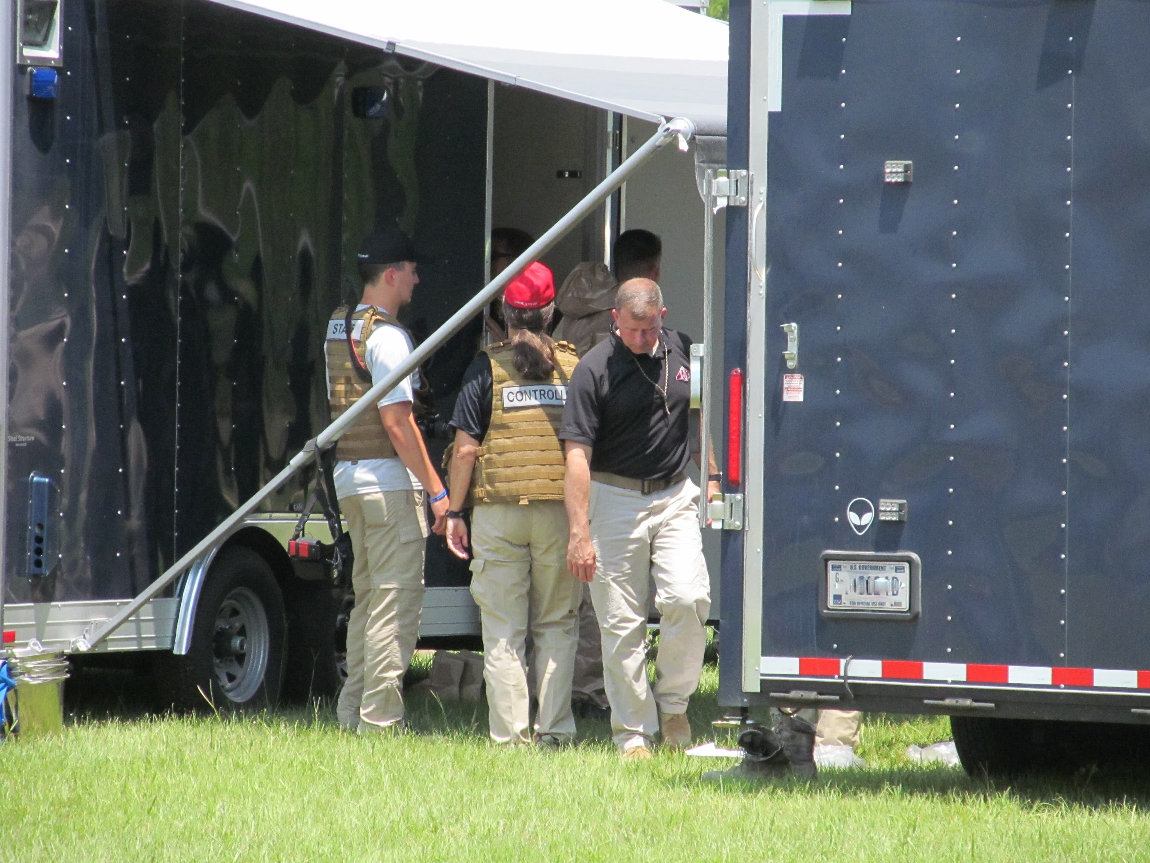 Instructors in vests and caps confer beside an open black equipment trailer on a lawn.