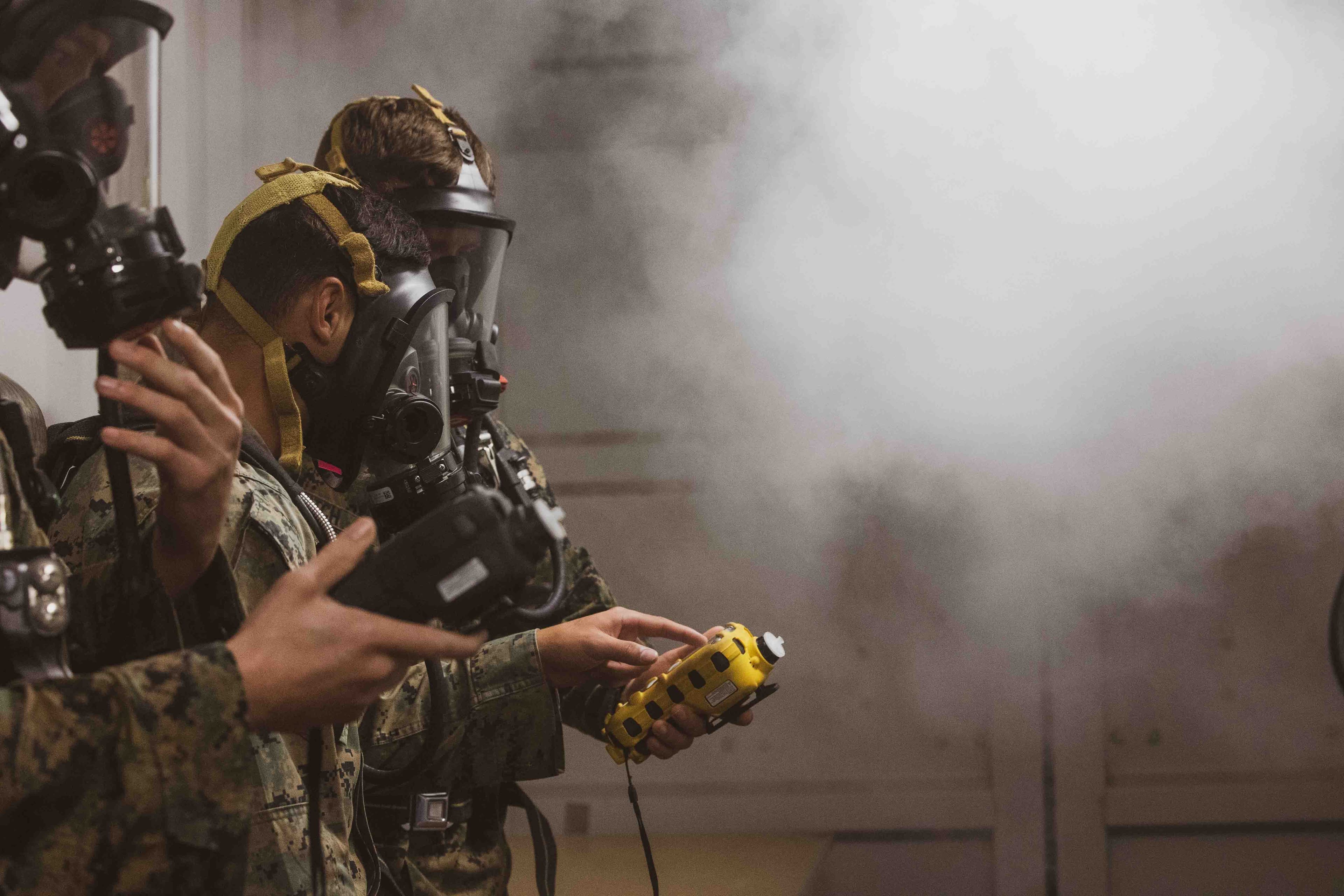 Marines in gas masks and camouflage handle a detector as smoke rolls through the room during CBRN training.