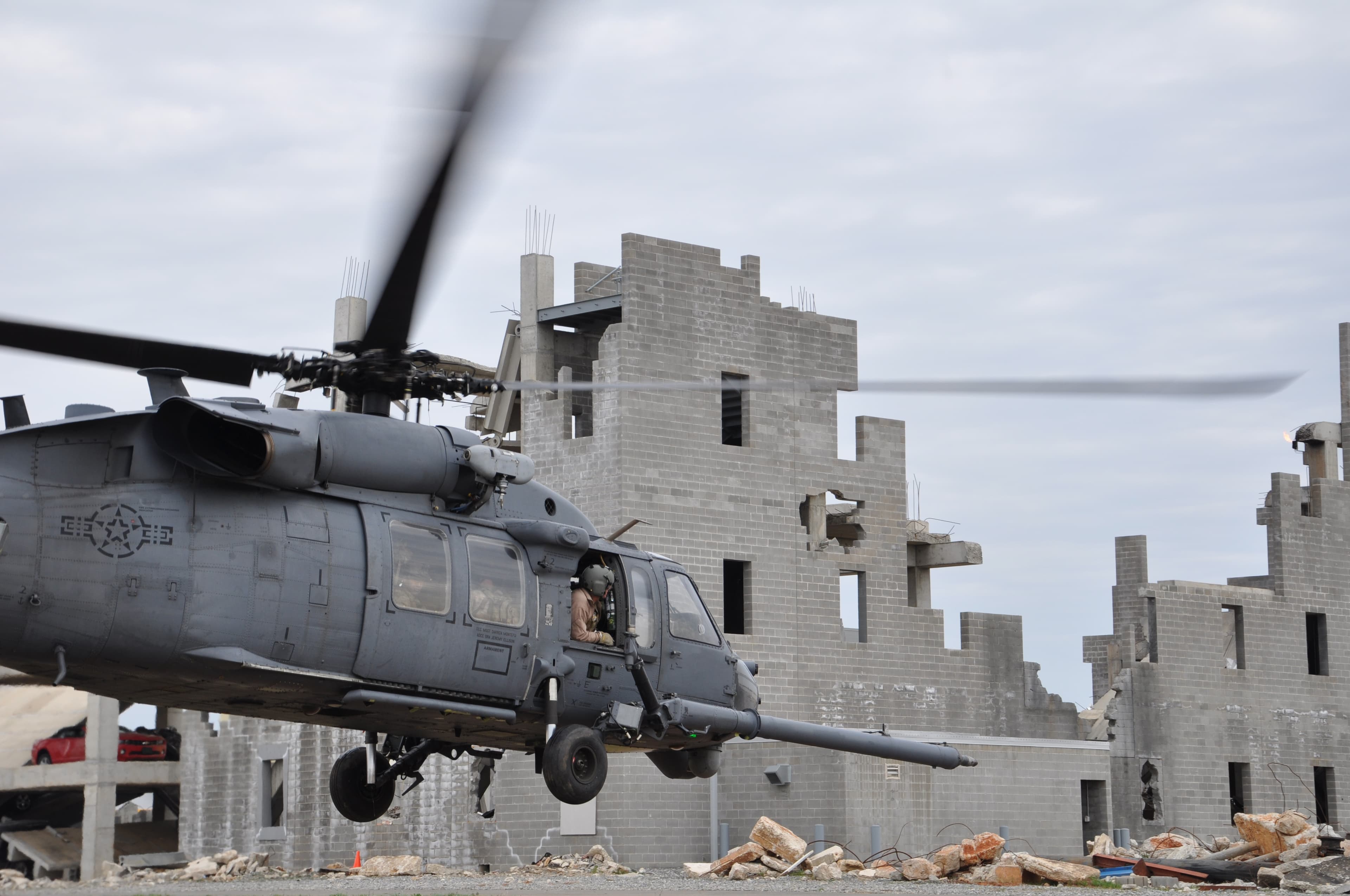 A military helicopter hovers low in front of a damaged concrete-block building, rotors blurred.
