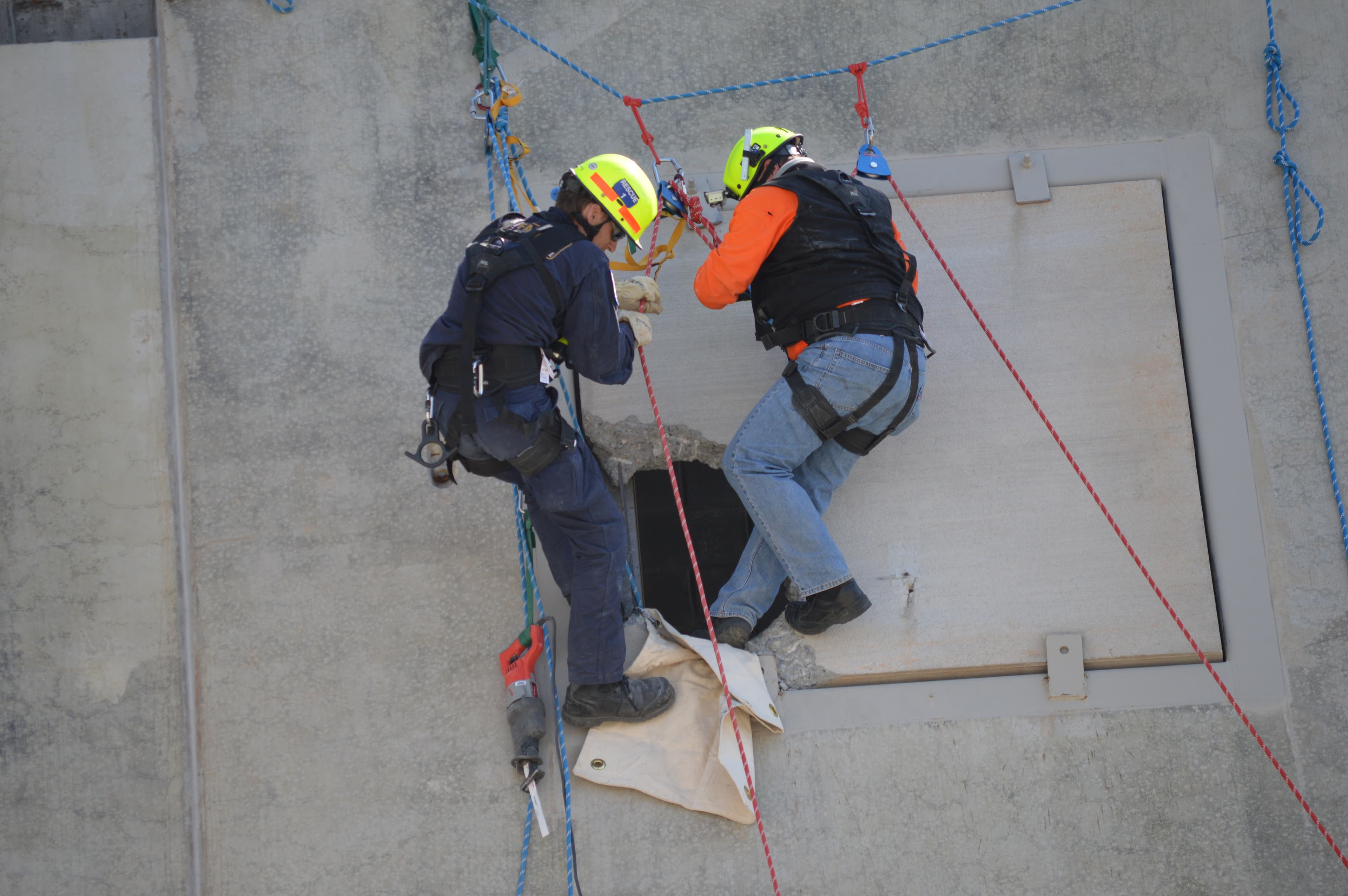 Two rescuers on ropes work at a hatch on a concrete wall, seen from above.
