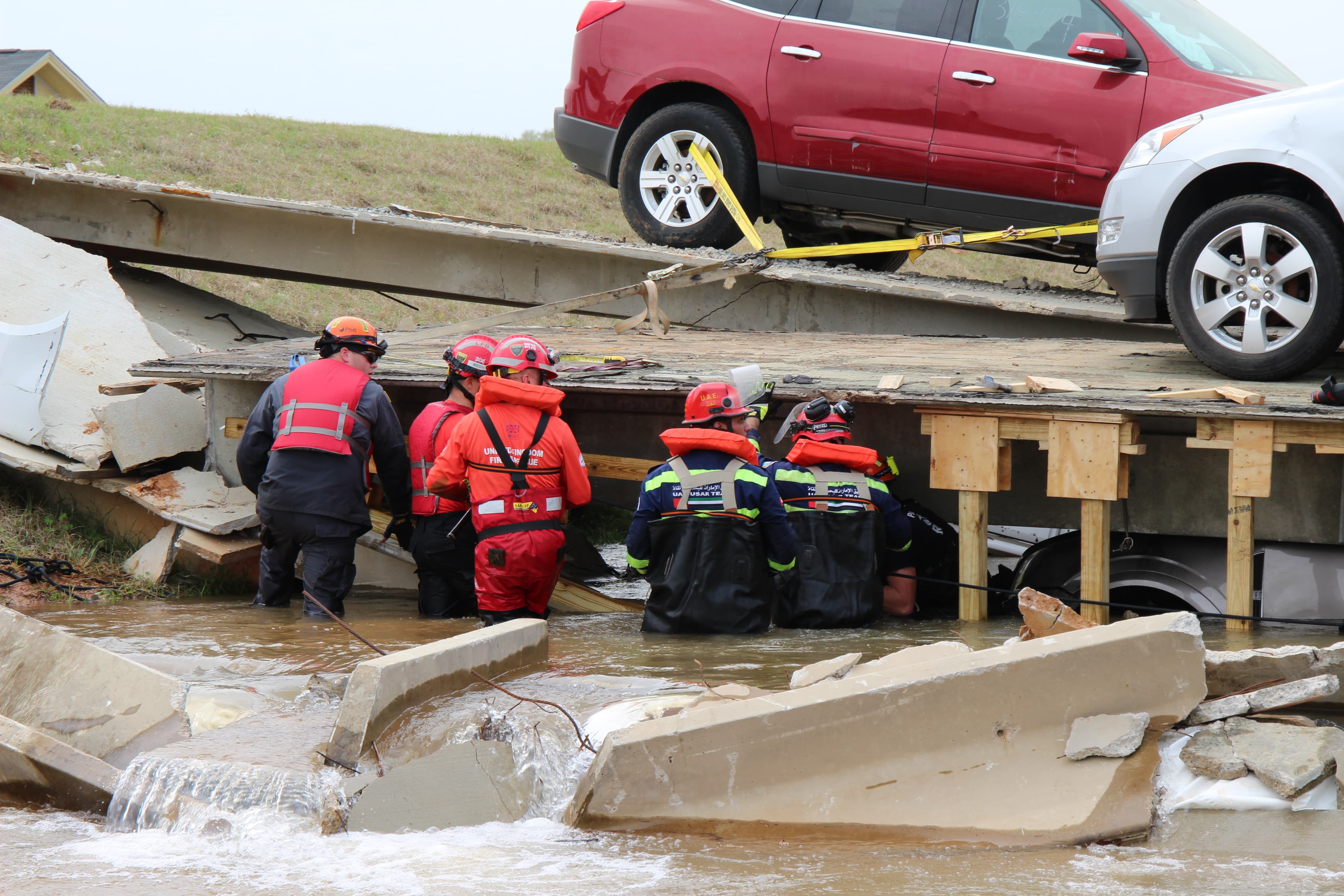 Rescuers in red life jackets wade through floodwater beneath a collapsed slab, with a car perched on the debris above.