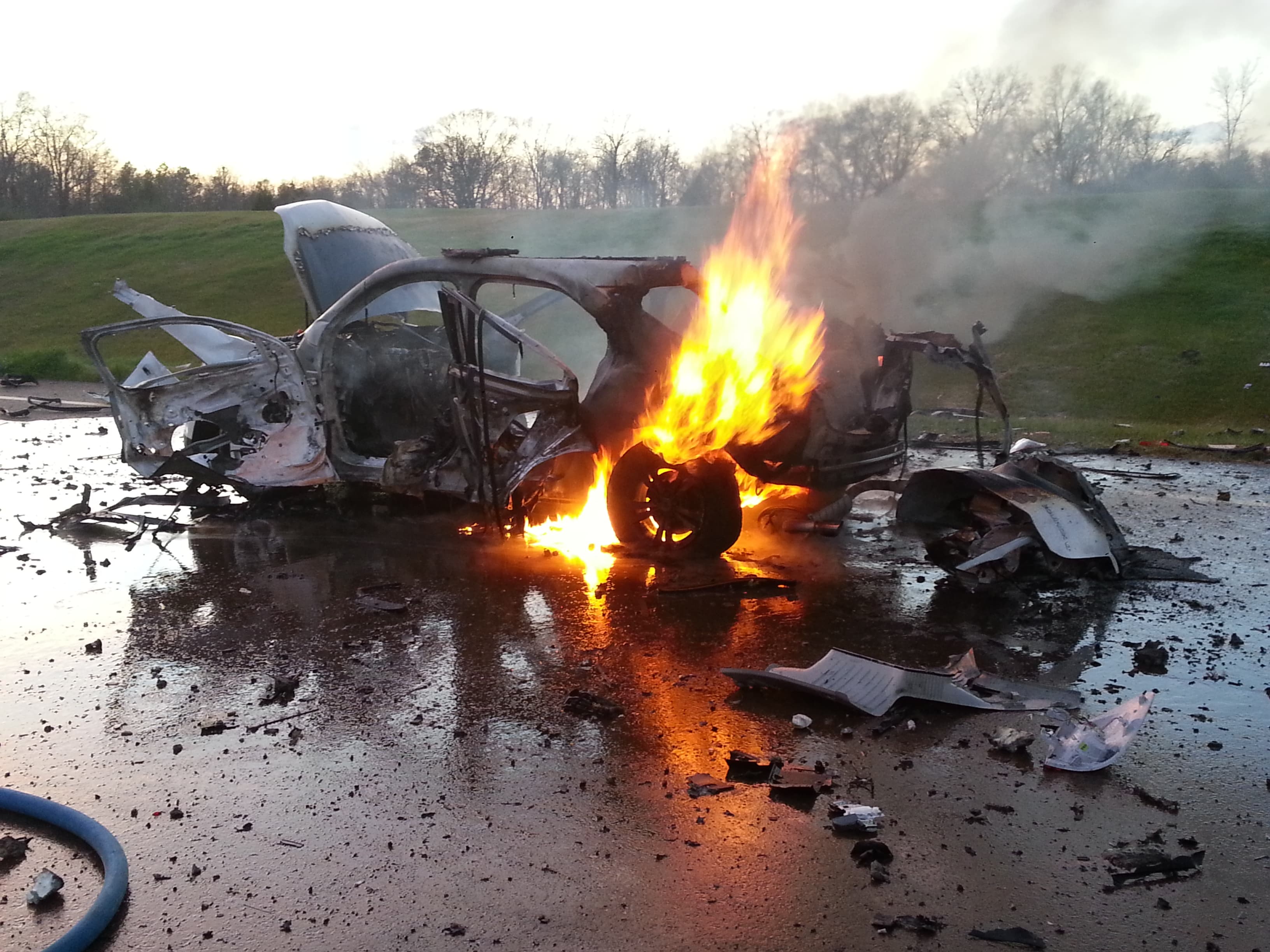 A car burns after an explosive demonstration, its shell torn open and debris scattered across the wet pavement.