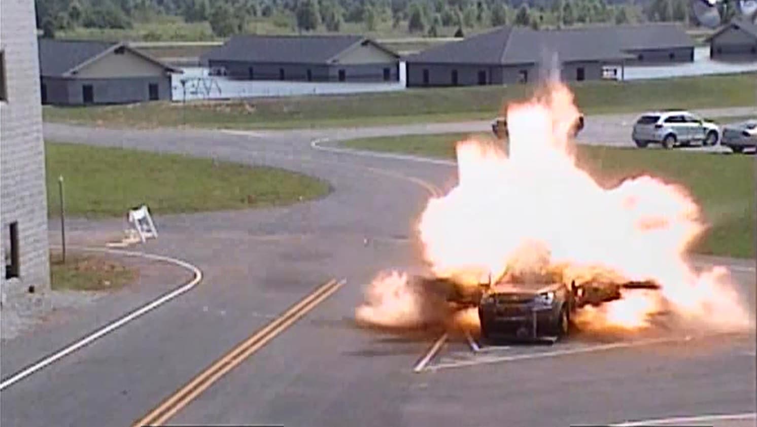 A parked car explodes in a burst of flame and smoke in front of the training buildings, seen from a camera.