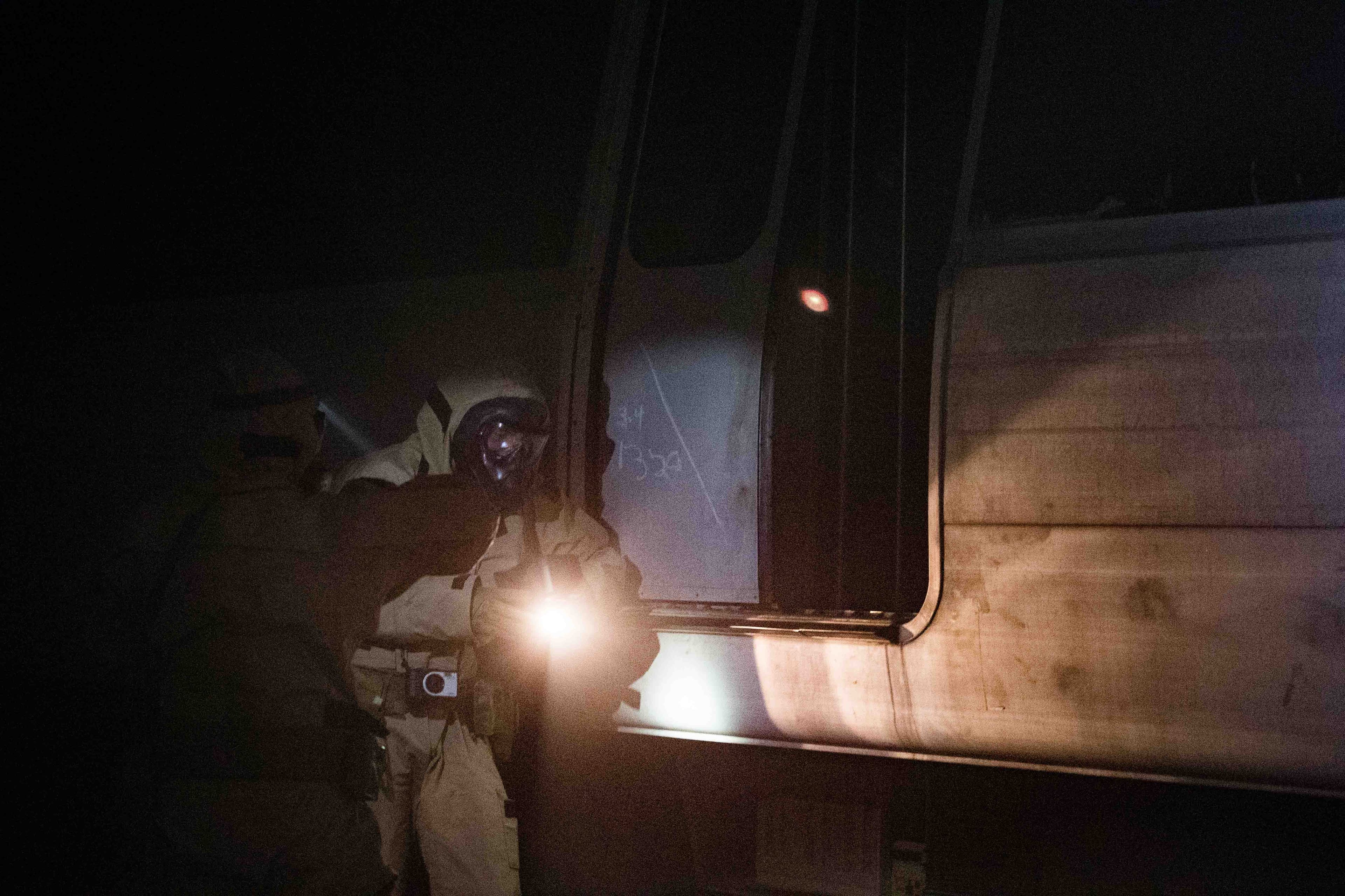 Two service members in desert camouflage and full-face respirators enter a darkened passenger rail carriage at night, one lighting the doorway with a handheld flashlight.