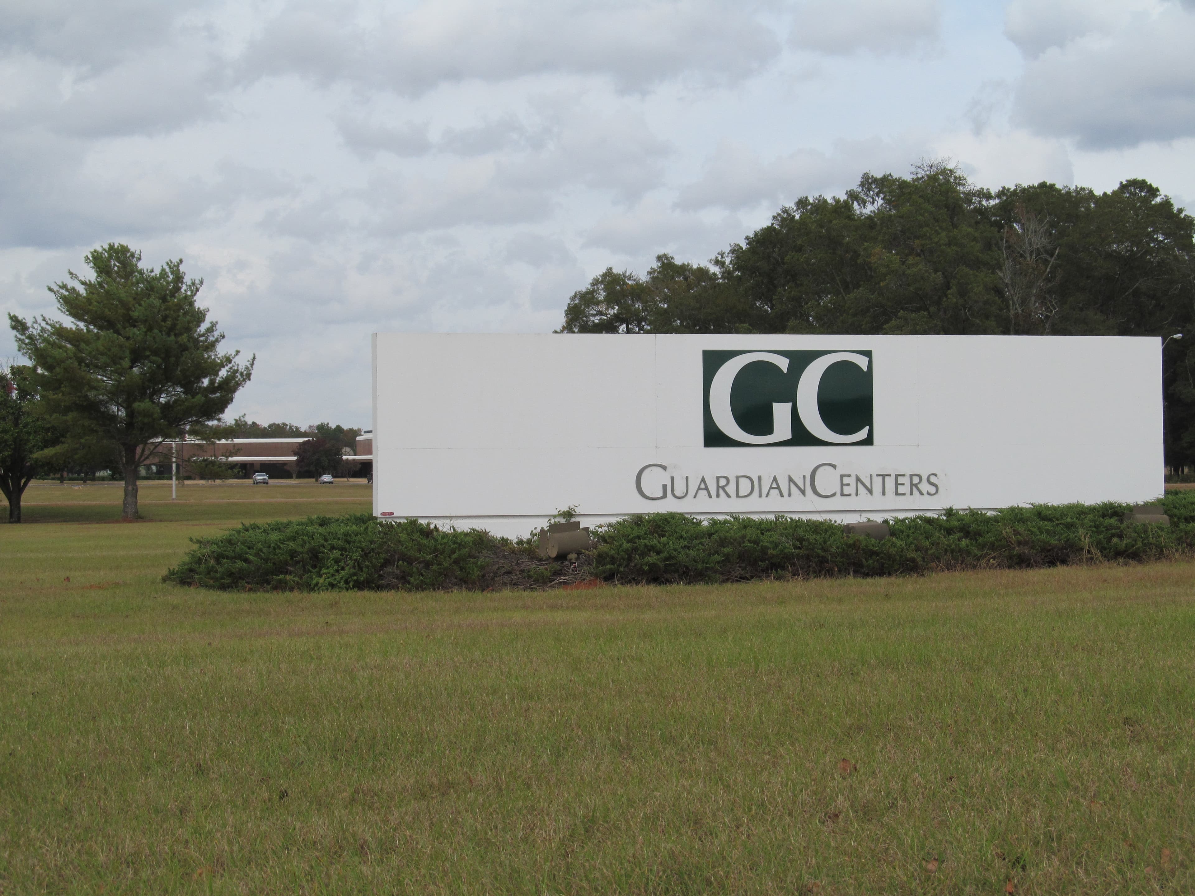 The Guardian Centers entrance sign, a white panel with the green GC logo, standing on a lawn in front of trees.