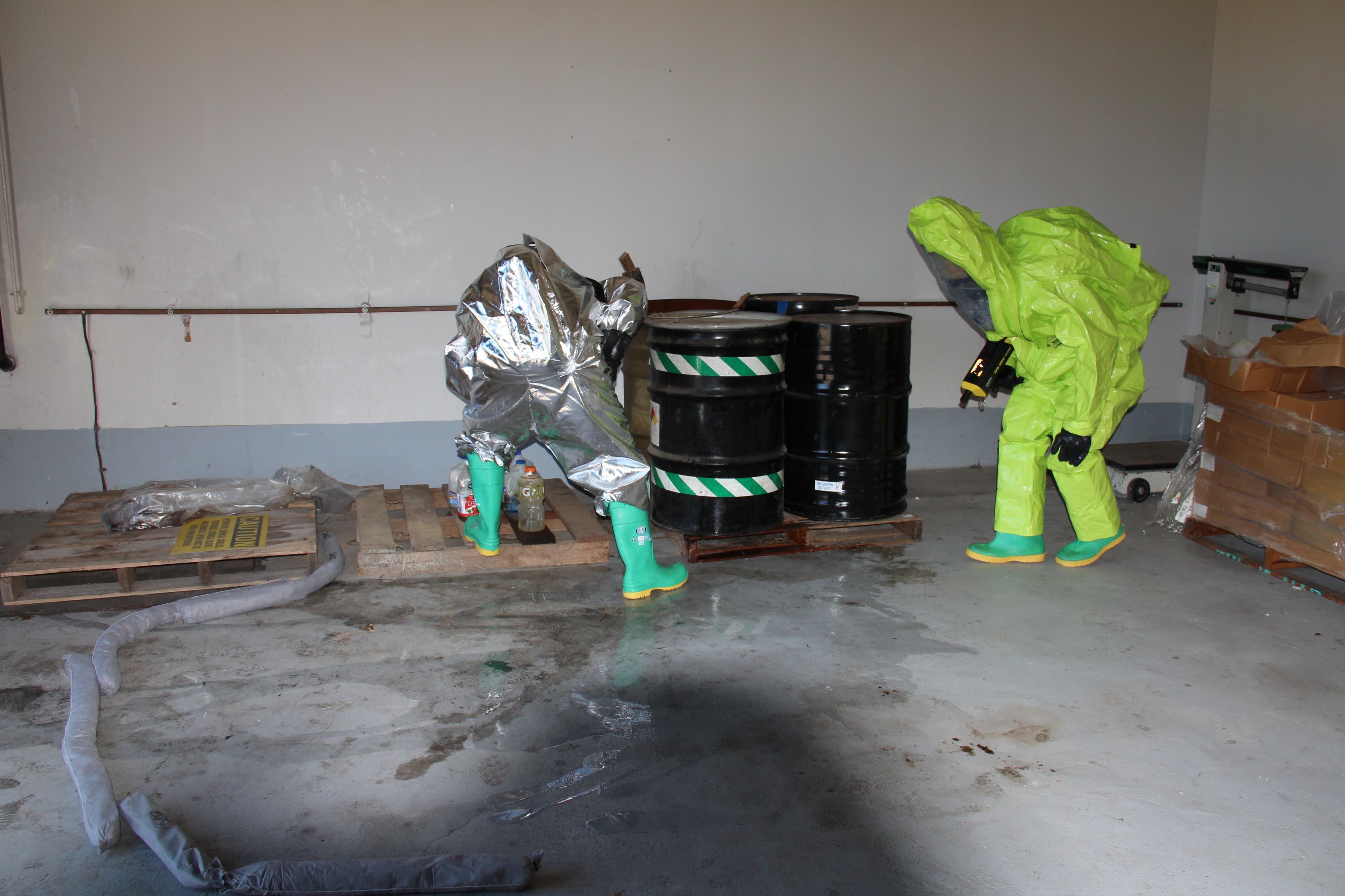 Two responders in silver and green hazmat suits inspect leaking drums inside a concrete storage room.