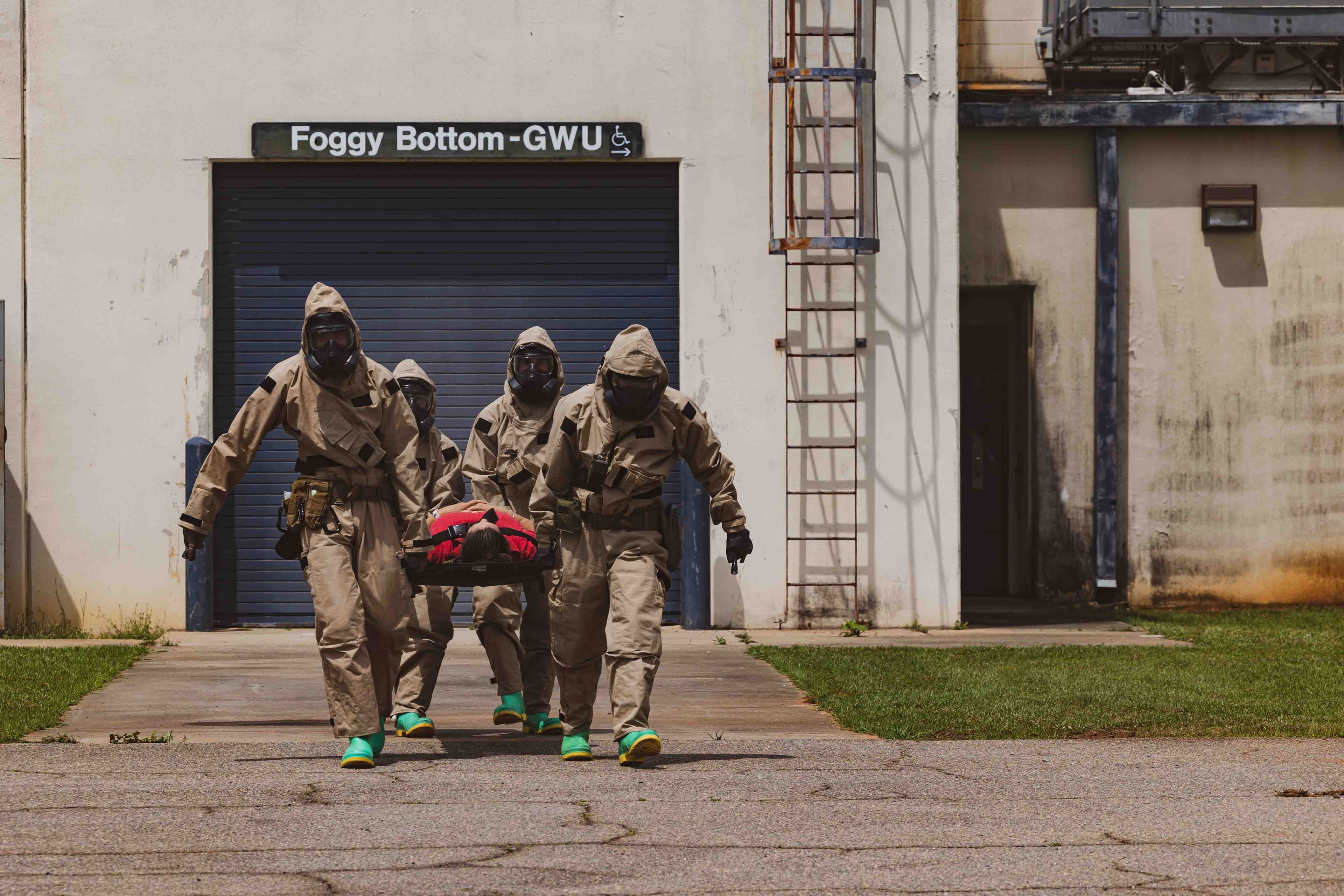 Marines in protective suits and gas masks carry a casualty on a litter past a subway station entrance marked Foggy Bottom-GWU.