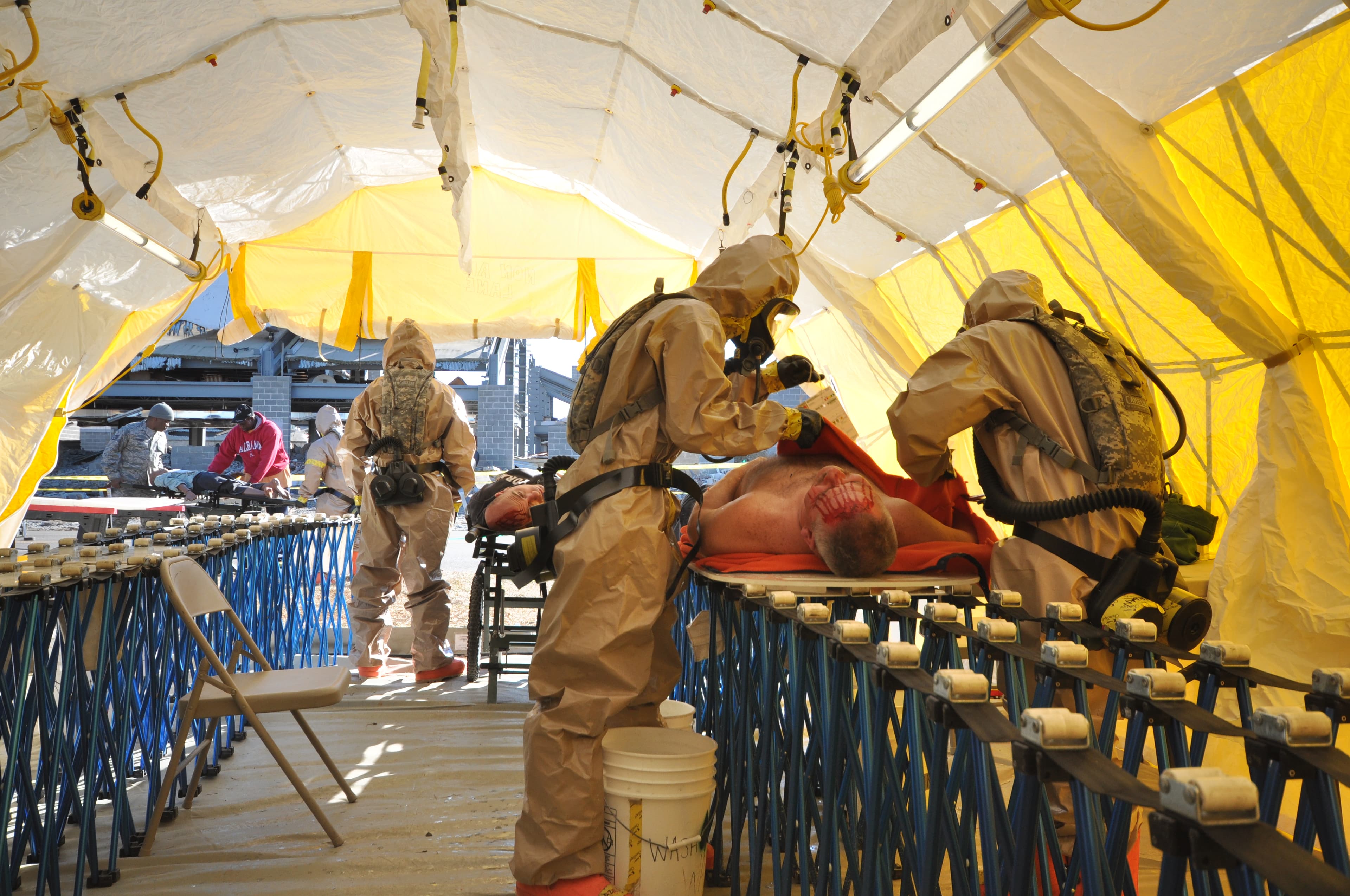 Responders in tan protective suits and masks treat a casualty on a stretcher inside a yellow decontamination tent.