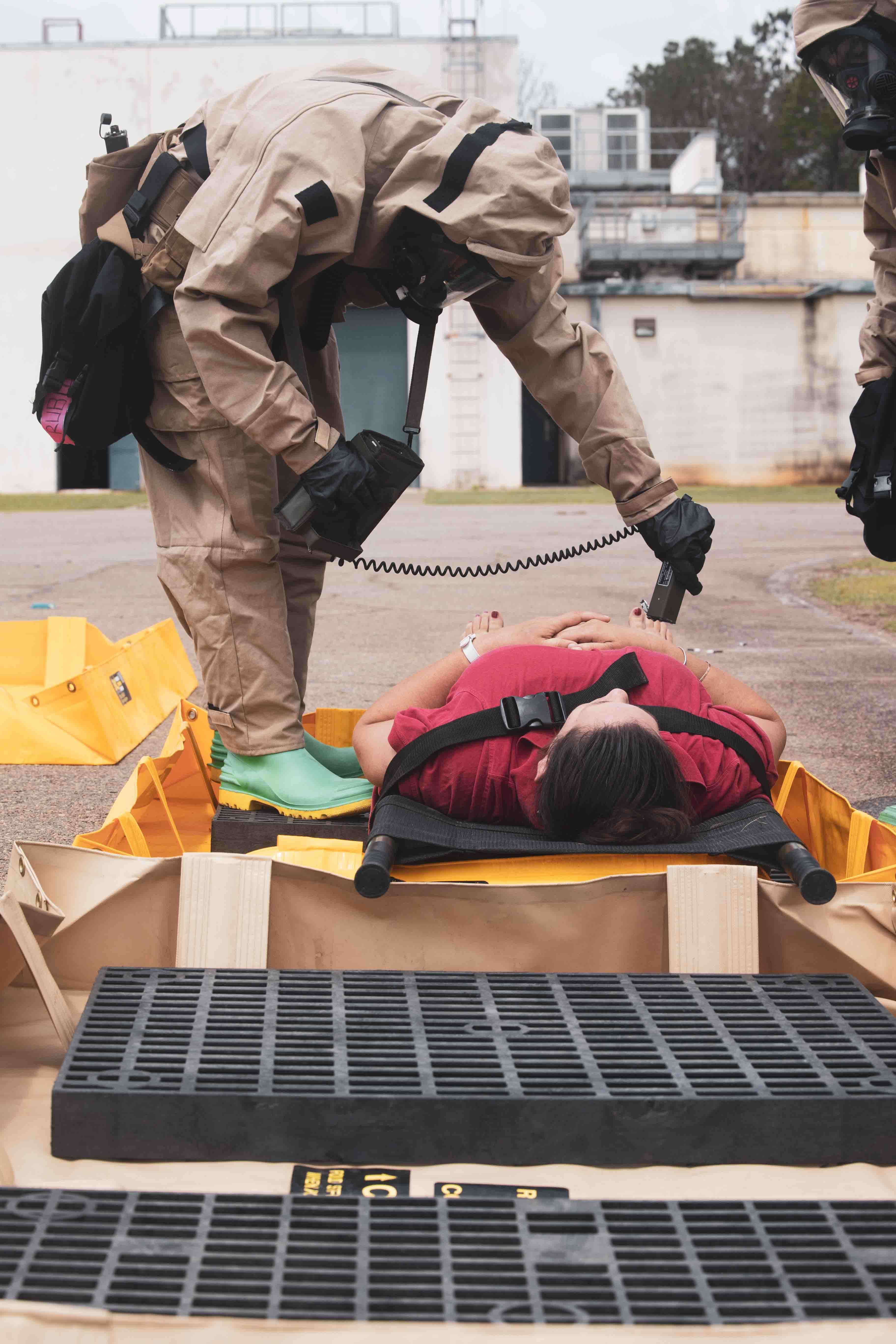 A Marine in a protective suit and mask uses a hand-held detector over a casualty lying on a decontamination board.