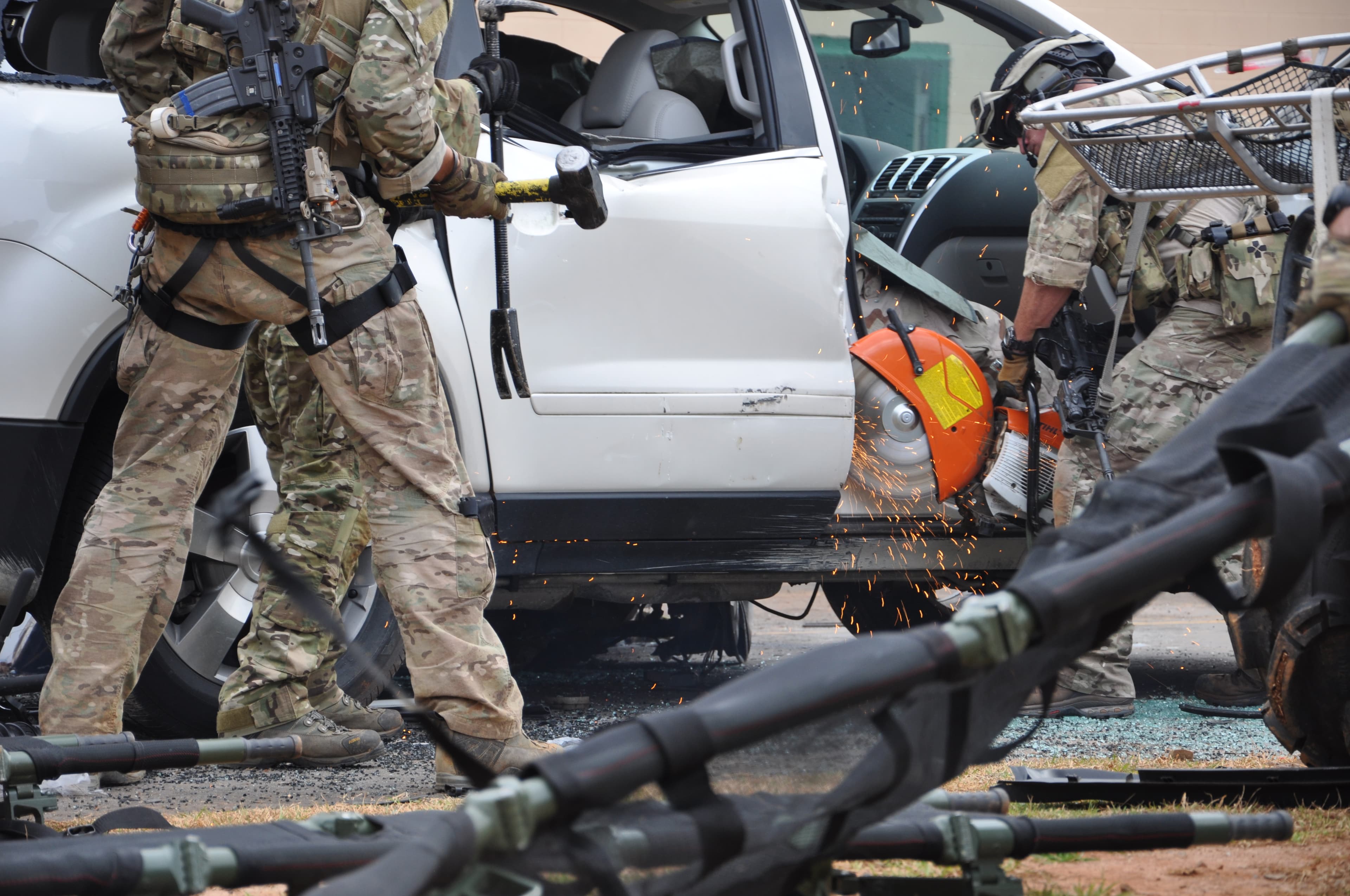 Soldiers cut open a white SUV with a rescue saw, sparks flying, in a vehicle extrication drill.