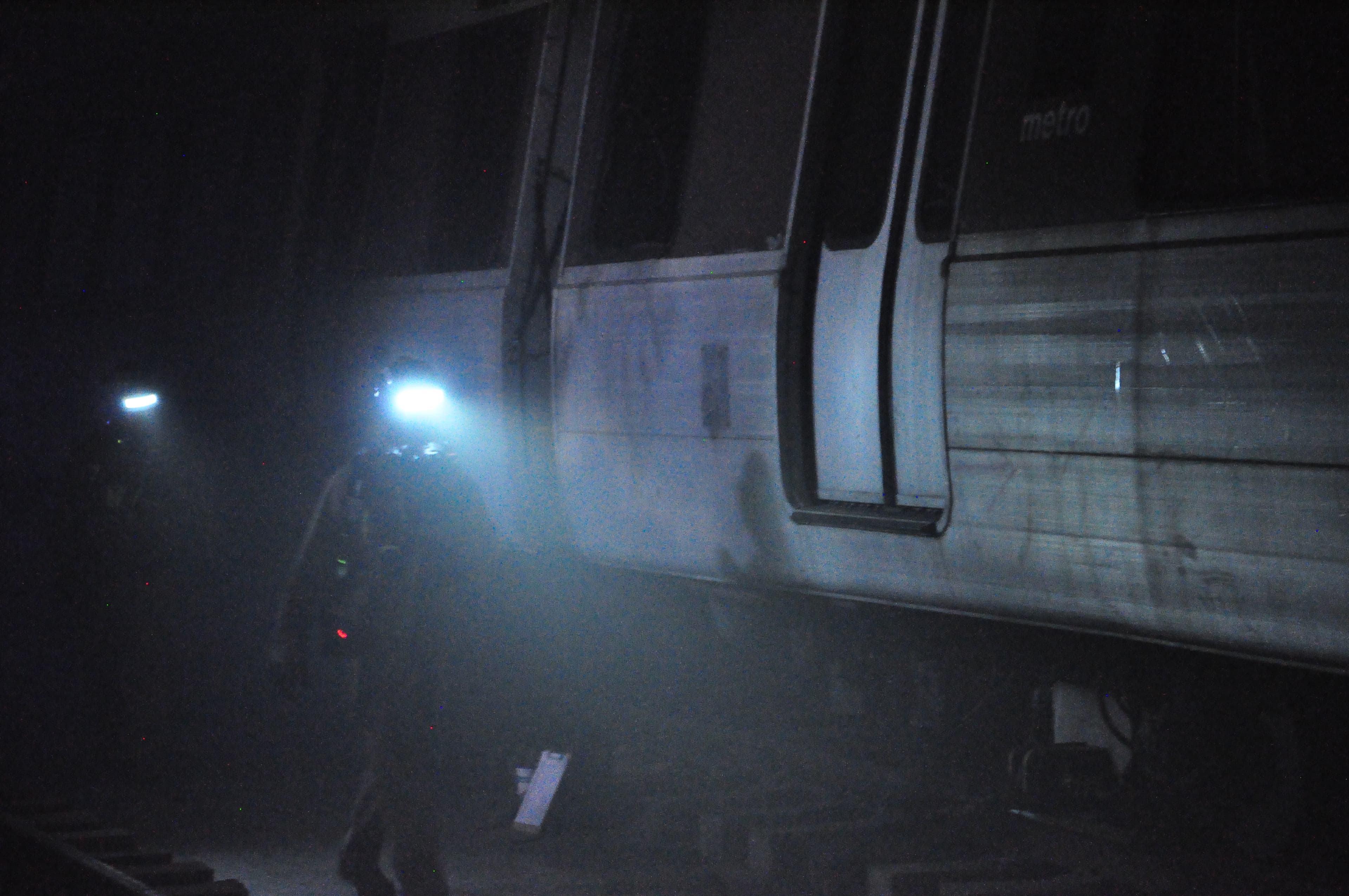 A rescuer's headlamp cuts through the darkness beside a subway car in the unlit rail tunnel.
