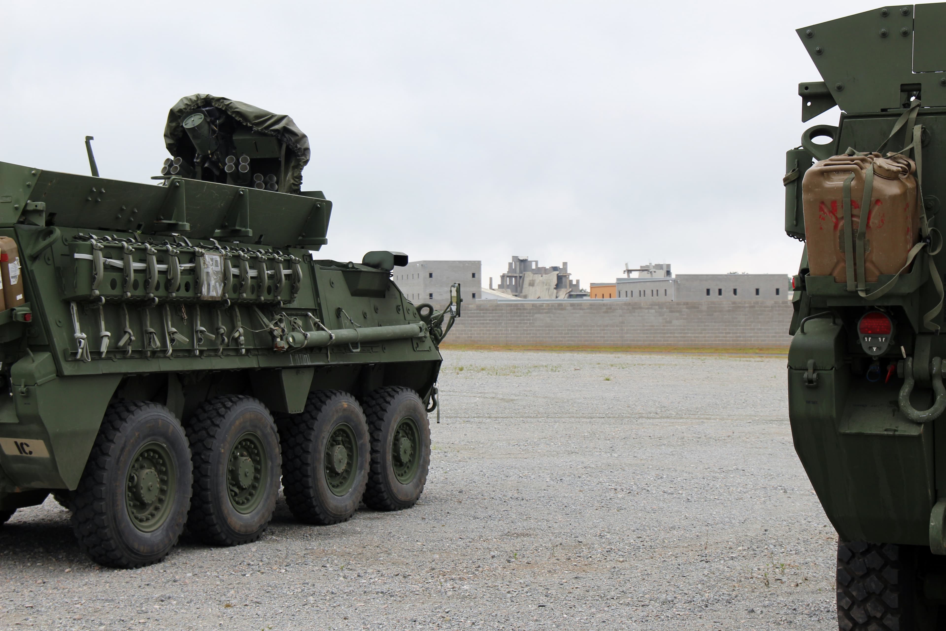 Armored military vehicles parked on a gravel staging lot with training buildings in the distance.