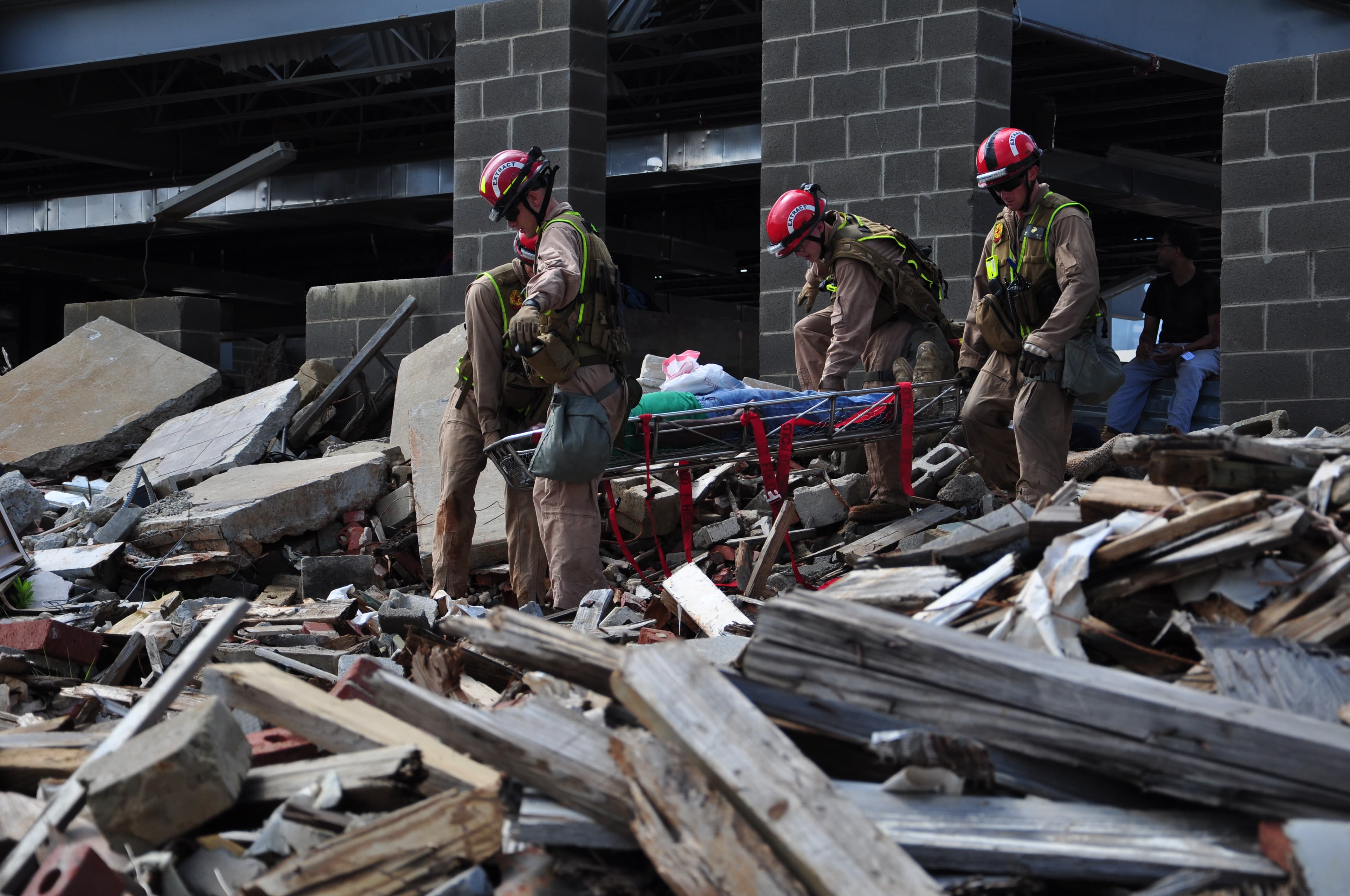 A rescue team in red helmets carries a stretcher across a field of collapsed-building rubble.