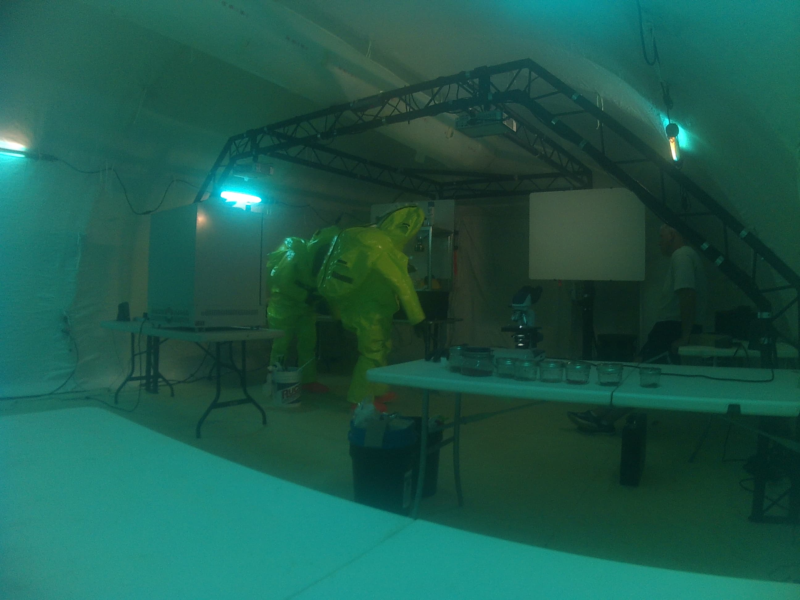 Inside a tent lit in green, a person in a yellow protective suit works at a table of equipment under a lighting truss.