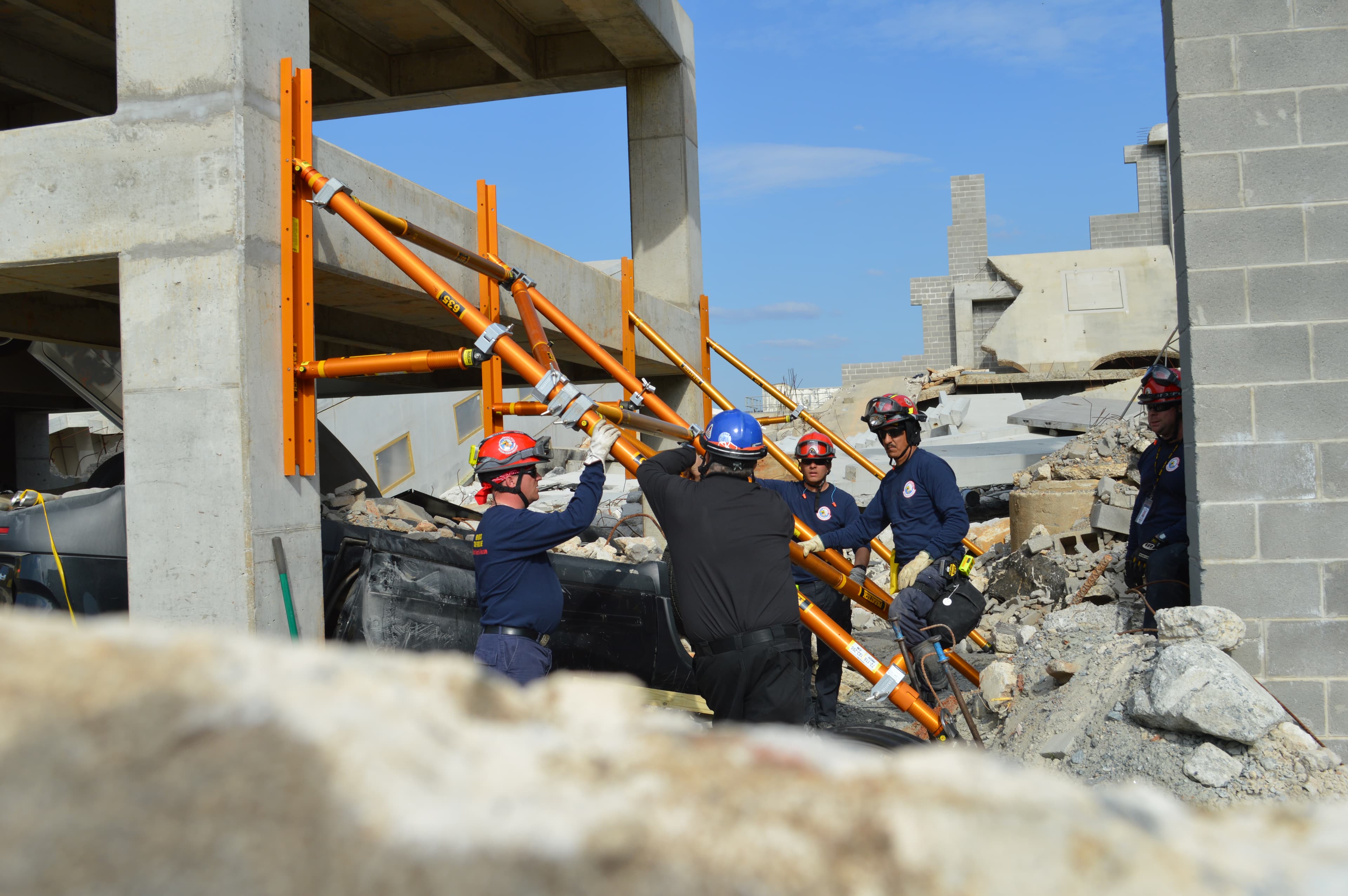 A rescue team raises orange shoring struts beneath a damaged concrete structure.