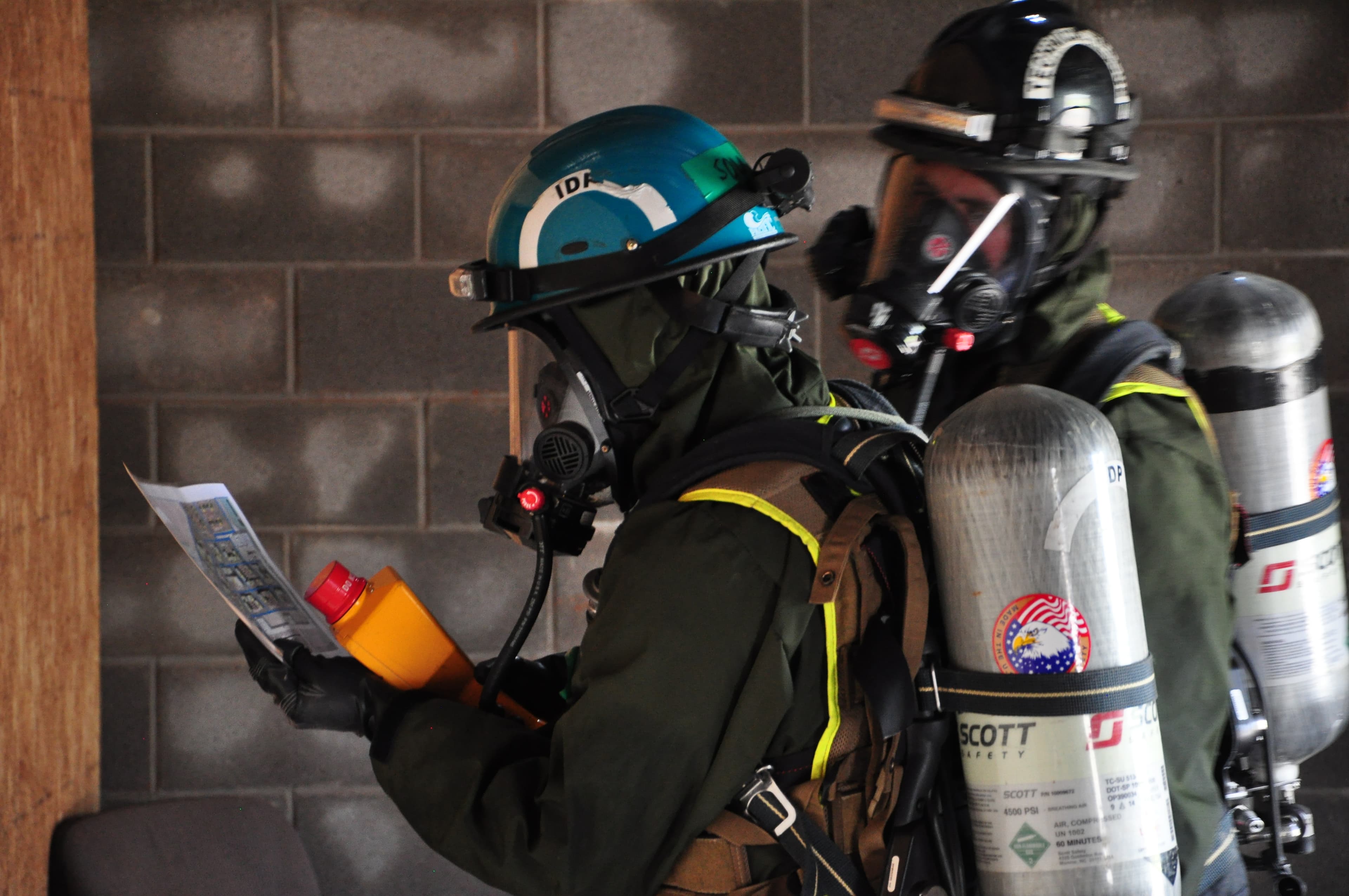 Two firefighters in breathing apparatus, one holding a radiation detector and a map, move through a concrete-block corridor.
