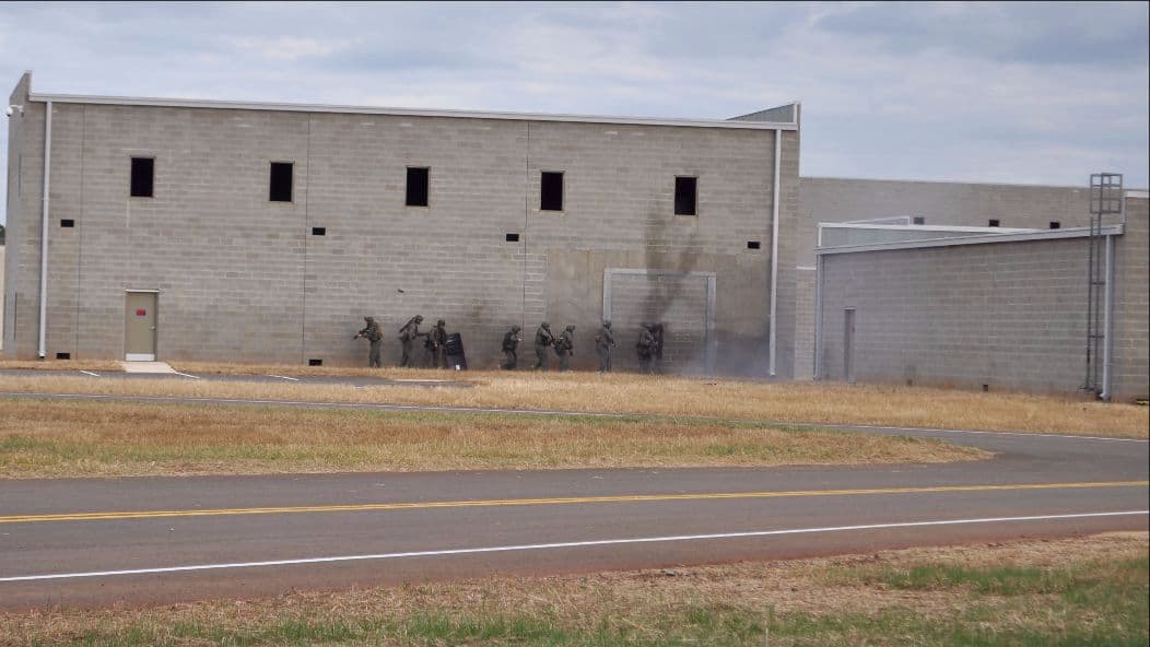 A tactical team stacks up at the door of a concrete-block training building as smoke rises from the entry.