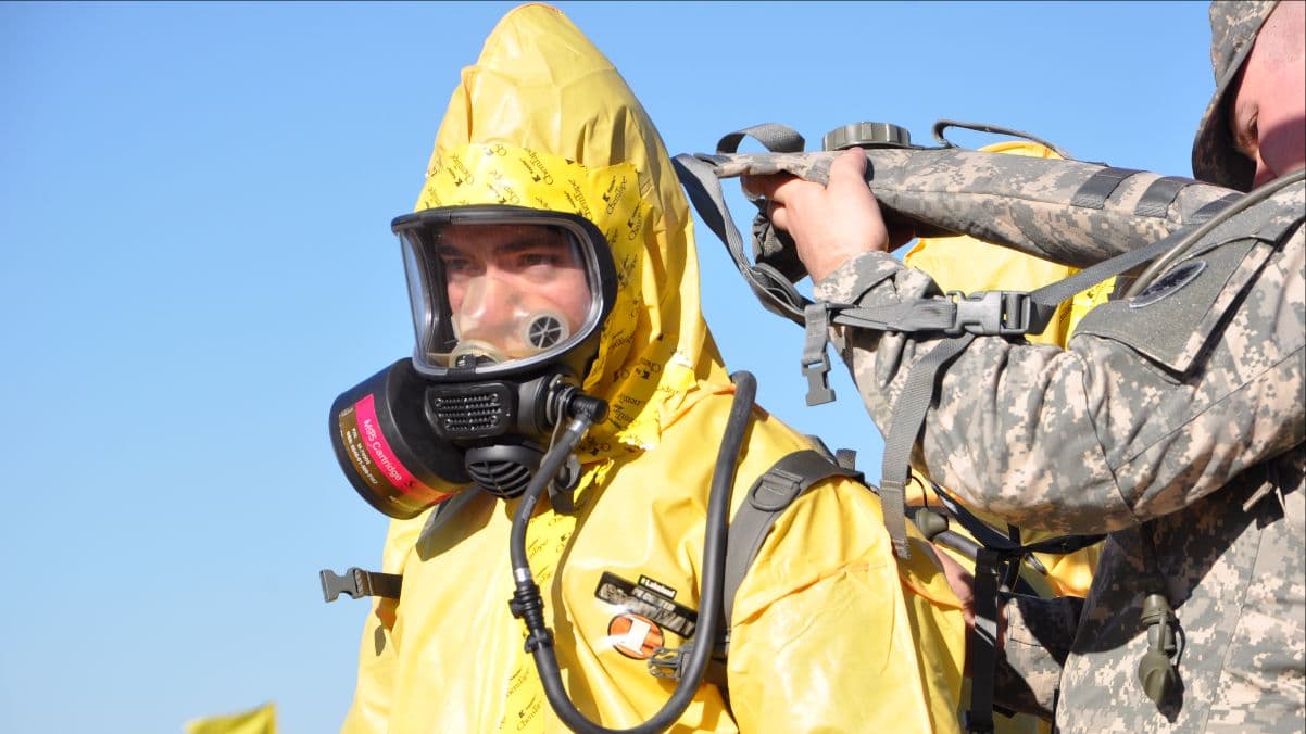 A soldier helps a trainee in a yellow hazmat suit and respirator fasten breathing apparatus.