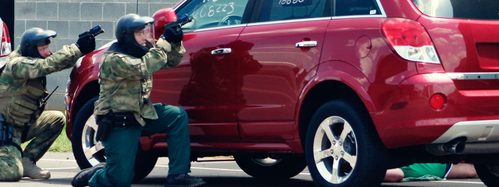Two officers in camouflage take cover behind a red SUV, aiming their weapons over the hood.