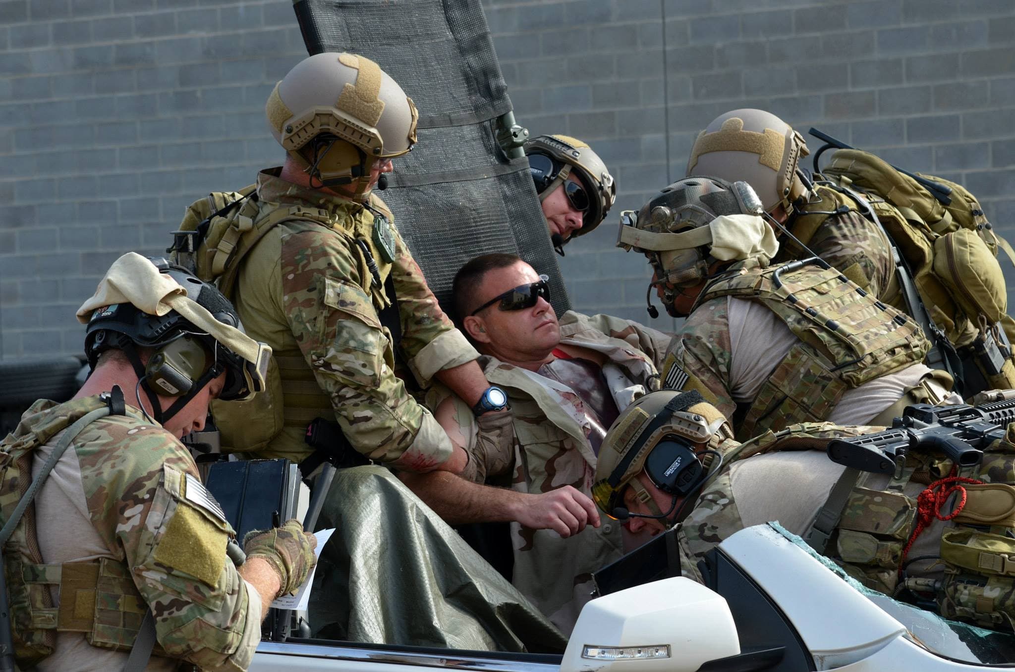 Soldiers in helmets and body armor treat a casualty on the hood of a white car.