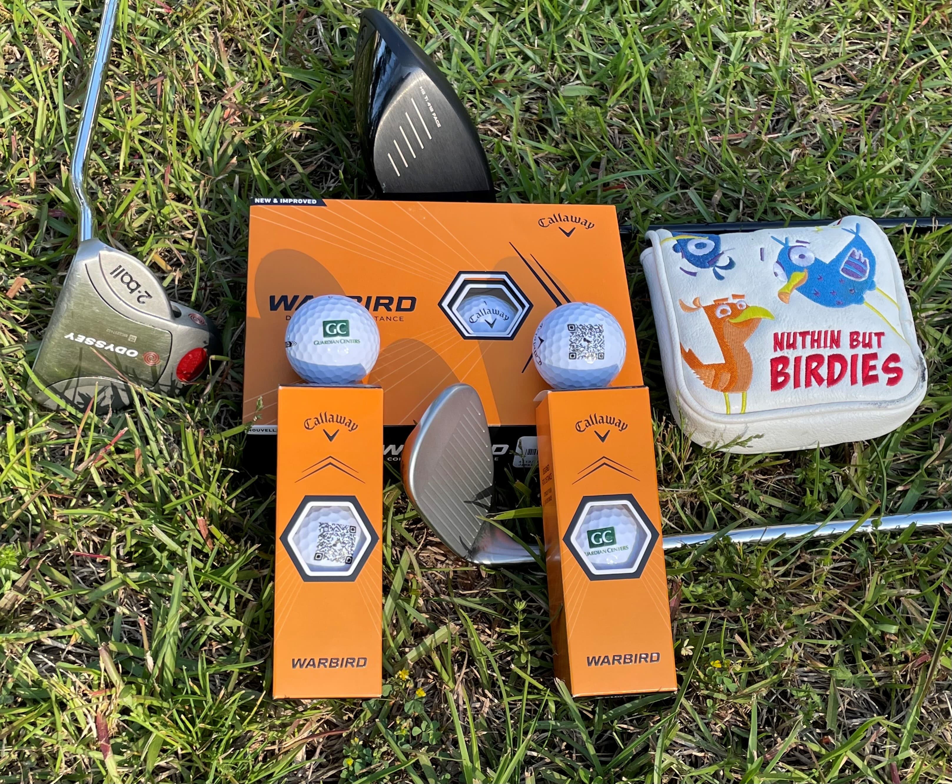 Golf clubs, orange Callaway Warbird ball boxes and a “Nuthin But Birdies” putter cover laid out on grass, with two custom balls — one printed with the Guardian Centers logo, one with a QR code.