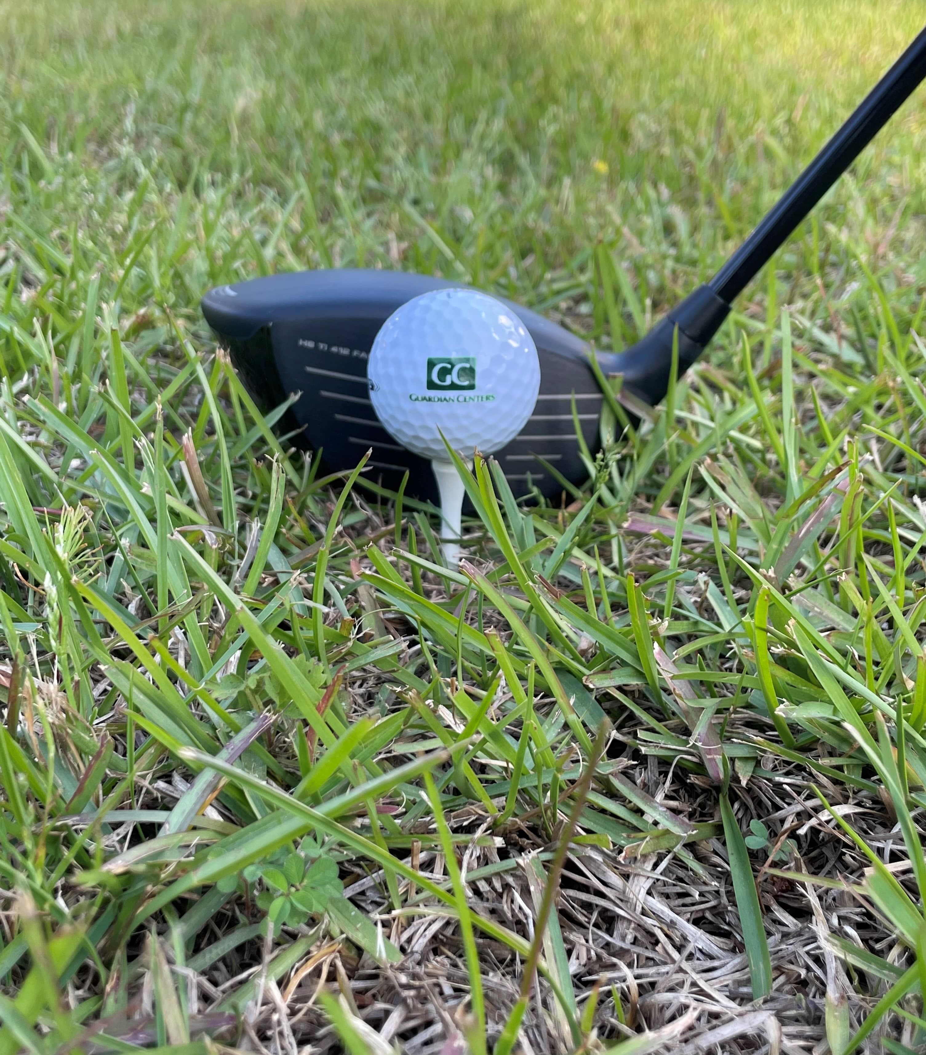A golf ball printed with the GC logo sits on a tee in the grass in front of a driver.