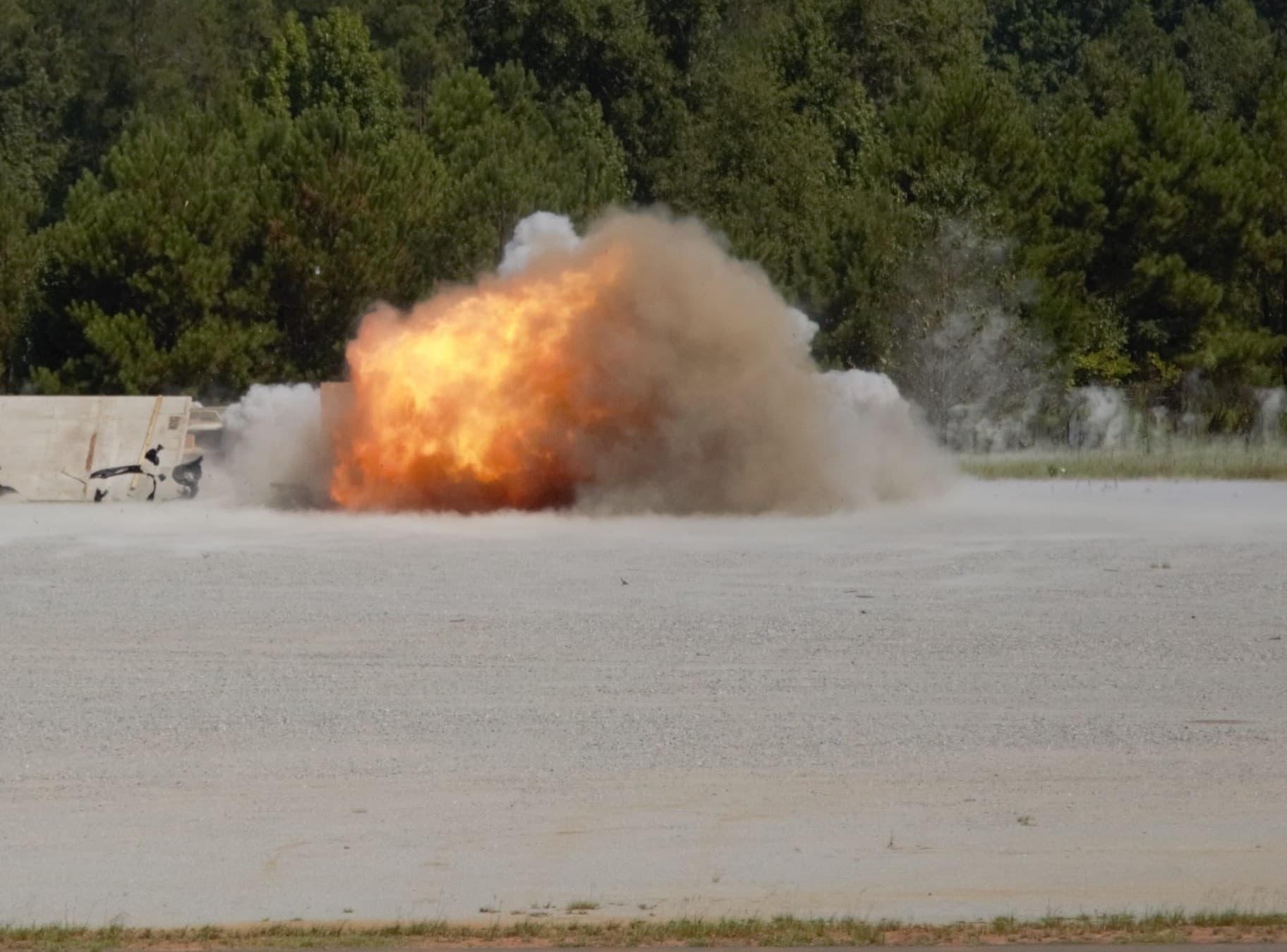 An explosive charge detonates in a fireball and dust cloud on the range in front of a treeline.