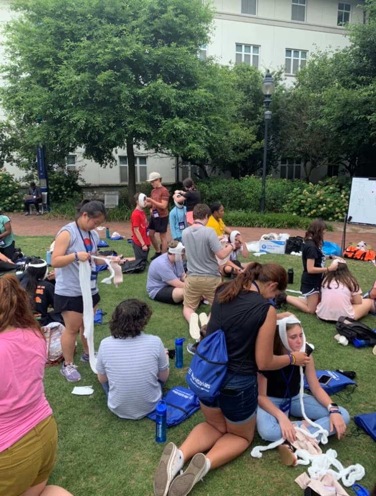 Students sit on a lawn practising bandaging on each other's arms during a first-aid session.