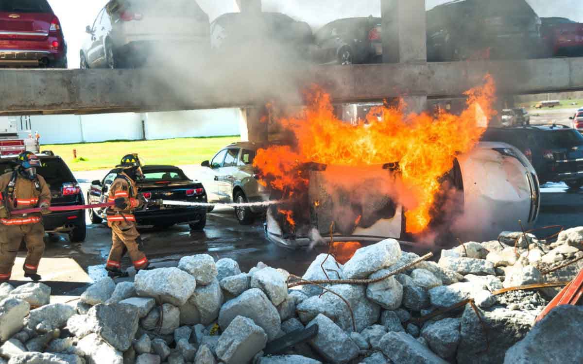 Firefighters in turnout gear advance on a burning vehicle beneath a collapsed parking deck, with rubble in the foreground.