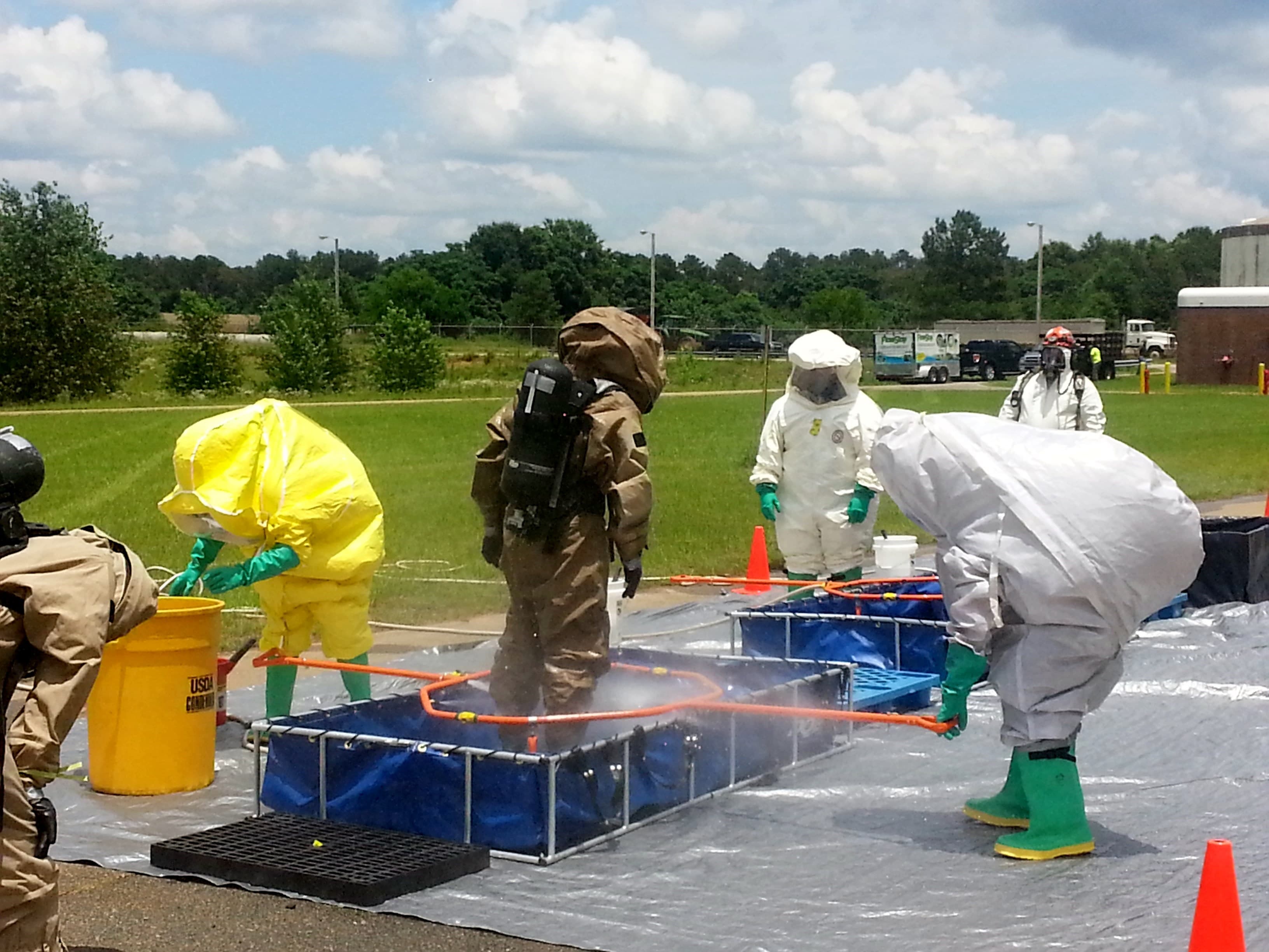 Responders in yellow, tan and white hazmat suits run a decontamination line of pools and sprayers on a lawn.