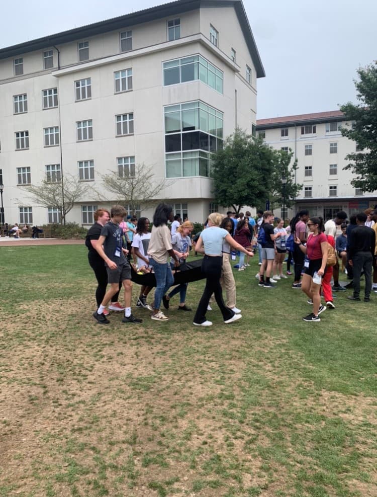 Students practice carrying a classmate on a stretcher across a lawn in front of a university building.
